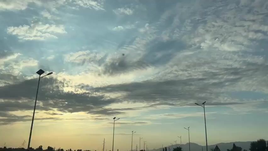 The video shows a moving shot of a sky with clouds and streetlights. The sky is a mix of blue and white, with some yellow and orange hues near the horizon, suggesting it is either sunrise or sunset. There are several streetlights visible, spaced along what appears to be a road. In the distance, there are silhouetted trees and hills. The clouds are varied, with some appearing wispy and others more dense. There is a noticeable dark smudge or spot in the sky in the upper middle portion of the frame that remains relatively consistent throughout the clip.