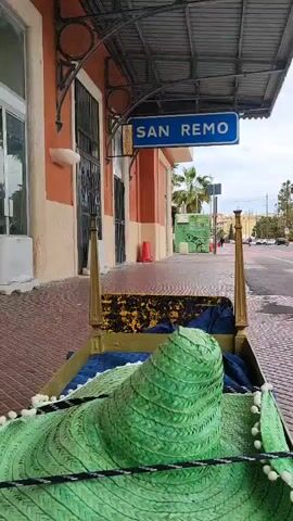 The scene features a bed on wheels positioned on the sidewalk in front of a building with a sign that says "SAN REMO". The bed frame is gold, and a large green sombrero with white pom-poms sits atop a blue blanket in the bed. The building is painted in muted orange and beige tones. The sidewalk is made of red bricks. Across the street, there's a graffiti-covered wall and some palm trees. In the distance, cars are visible on the road. The sky is overcast. Sounds of music can be heard in the background.