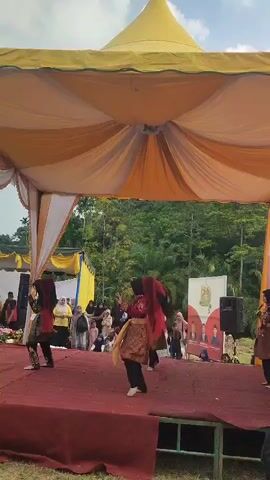 A group of four female dancers in traditional attire are performing on a red carpeted stage under a yellow and white striped tent. The dancers, wearing headscarves and ornate clothing, are in the midst of a coordinated dance. Behind the stage, a crowd of spectators are gathered, some standing and some seated. In the background, lush green trees and foliage are visible under a partly cloudy sky. A banner featuring several men in suits hangs on a white structure to the right of the stage, and a speaker is positioned on the right side of the stage. The scene appears to be an outdoor event or festival.