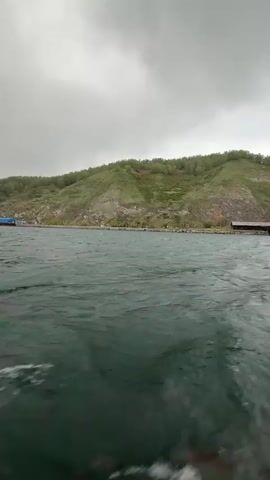 The video shows a view from a boat on a body of water. In the background is a large hill covered in green vegetation and some exposed rock. The sky is overcast. There's also a dock or other structure visible in the distance. The water is dark and choppy, indicating some wind.
