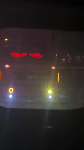 The video shows a car's rear window at night. The window is somewhat dirty or wet, obscuring the view of the streetlights behind it.  The main focus is on the red brake lights that are illuminated in the shape of two horizontal, curved, narrow bars, similar to the shape of stylized eyes.  There are also other lights visible through the window, including yellow and white streetlights and vertical red lights. The camera is relatively still while focused on the lights. The overall impression is that the video was taken from inside a parked car, looking out at night.