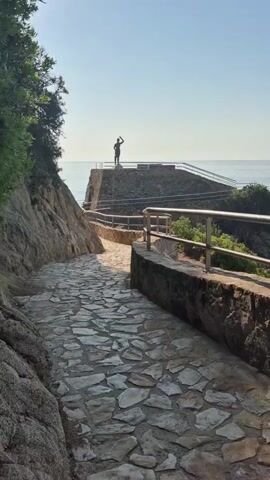 A person stands on a rocky outcrop overlooking the sea, raising their hand in a gesture of greeting or farewell. A stone pathway winds its way up to the outcrop, bordered by a stone wall and a metal railing. Lush green foliage grows on the left side of the pathway, contrasting with the rough texture of the rocks. The sky is a clear blue, and the sun casts a warm glow over the scene, suggesting a peaceful, scenic location.