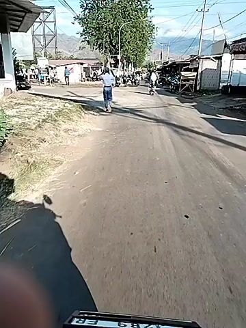The video begins by showing a man in a white shirt and blue jeans standing on a paved road, facing away from the camera. He appears to be interacting with someone off-camera. The road is flanked by buildings and stalls, with several motorcycles parked along the side. In the background, mountains are visible under a clear sky. As the video progresses, more motorcycles and people come into view, suggesting a busy marketplace or gathering. The camera, seemingly mounted on a motorcycle, moves forward, capturing the scene from a first-person perspective. The road surface is uneven with some potholes. The overall impression is a lively street scene in what appears to be a Southeast Asian or similar environment.