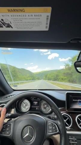 The video shows a first-person perspective from inside a car, driving on a highway. The driver's hands are on the steering wheel of a Mercedes-Benz. The dashboard displays speed and other information. Through the windshield, a road stretches ahead, flanked by green, tree-covered hills under a blue sky with white clouds. The sun is shining. A warning label about advanced airbags is visible on the car's visor.