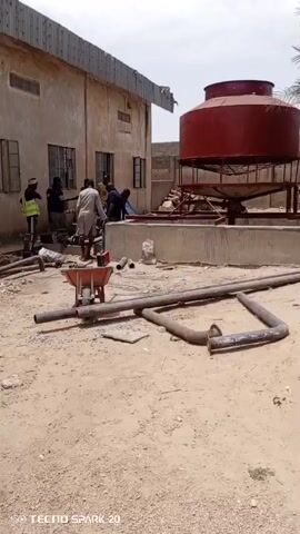 The video shows a group of men working near a large red water tank on a concrete base. The tank is located next to a beige building with several windows and a corrugated metal roof. Pipes are scattered on the ground in front of the tank, along with a small red wheelbarrow filled with tools. The men are dressed in various casual clothes, and one is wearing a high-visibility vest. They appear to be engaged in some kind of construction or maintenance work, possibly related to the water tank or the building's plumbing. The ground is sandy, and the sky is overcast. A palm tree is visible in the background. The camera is stationary and captures the scene from a medium distance. A watermark "Tecno Spark 20" is visible at the bottom left corner.