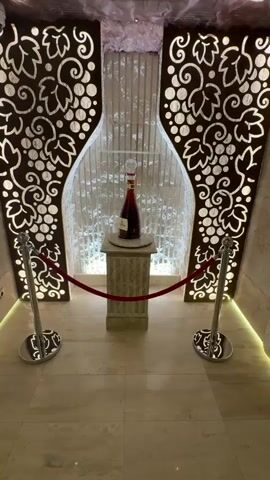 The video shows a display of a large, decorative bottle of liquor. The bottle is on a marble pedestal in the center of the frame. The pedestal is surrounded by velvet ropes and stanchions, suggesting the bottle is an exhibit. The background features a wall with a textured pattern and decorative panels on either side that resemble grape leaves. The lighting is dim, with spotlights highlighting the bottle. There are no people visible in the video.