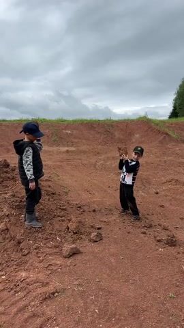Two young boys are outdoors in a large, reddish-brown dirt field under an overcast sky. The boy on the left, wearing a blue cap, black vest, and camouflage shirt, extends his right arm, and a dark clod of dirt is visible mid-air, moving from his left to the right side of the frame. The boy on the right, smaller and wearing a black cap and a black-and-white patterned long-sleeve shirt, is holding a large clump of reddish-brown dirt with both hands and takes a step. The background consists of sculpted dirt mounds, a distant line of green vegetation, and trees.