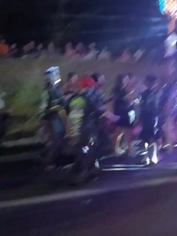 The video depicts an outdoor scene at night, with a crowd of people gathered on a grassy embankment overlooking a road. The lighting suggests artificial light sources, possibly from vehicles or event lighting, casting shadows and illuminating the faces of some individuals. Several people are seen on the road, some appearing to be interacting with each other, while others are observing the scene. The individuals on the embankment are mostly blurred due to the motion and lighting conditions, but they appear to be a mix of adults and possibly children. The overall atmosphere seems to be that of a public gathering or event, possibly a race or a performance, given the presence of a crowd and the organized arrangement of people. The video is shaky and the focus is inconsistent, indicating it was likely recorded handheld and possibly in a dynamic environment.