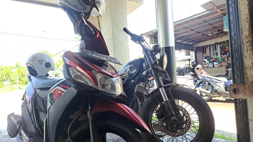 The image captures a roadside scene, likely the front of a small shop or garage, featuring two prominent motorcycles parked side-by-side under an overhang. In the foreground, a red and black automatic scooter is parked on the left. A gray helmet rests on its seat, and another, lighter-colored helmet hangs from its handlebar. The scooter's front headlight and fender are clearly visible, along with a partially obscured Indonesian license plate reading "KB 5886 B", indicating West Kalimantan.

Next to the scooter, on the right, is a black custom motorcycle, possibly a bobber or chopper, characterized by its spoked wheels and a large front tire. Its circular headlight and handlebars are visible, and a word that appears to be "Custom" or "Dubdee" is partially discernible on its fuel tank.

In the background, further along the right, several other scooters or motorcycles are parked, suggesting a wider parking area or a collection of bikes at a workshop. The background also reveals part of a building with an open front and a corrugated metal roof, where various tools or items are displayed or stored, including a hanging birdcage.

The scene is brightly lit by natural daylight, with a clear blue sky dotted with white clouds visible above the overhang. Green foliage from trees lines the distant road, and utility poles with electrical wires crisscross the sky. The sunlight creates shadows, suggesting a time between mid-morning and early afternoon. The setting, with its specific vehicle types and building architecture, is typical of a street scene in Southeast Asia, aligning with the provided location context of Mempawah, Indonesia. No people are visible in the frame.