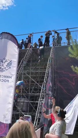 The video shows an outdoor obstacle course with participants climbing a tall, multi-tiered structure made of scaffolding and netting. People are seen actively climbing the structure using the netting and metal rungs. Several individuals in matching blue and white shirts appear to be part of a team or event support, standing near the base of the structure. Other people are watching from the ground, some holding cameras or phones. Large flags with logos are present, one prominently displaying a double-headed eagle emblem. The environment appears to be a sunny day with a clear blue sky.