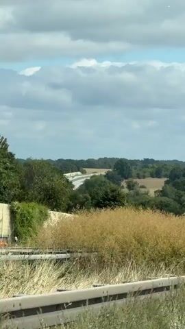 The video shows a highway with several trucks and cars traveling on it. The highway is surrounded by trees and fields. The sky is cloudy. The camera appears to be mounted on a vehicle moving alongside the highway. The video is shot from a slightly elevated perspective. In the foreground, there is tall, dry grass and a metal guardrail. In the middle ground, the highway curves to the left, with vehicles traveling in both directions. Several trucks are visible, some of them bright yellow. In the background, there are more trees and hills. The overall impression is of a rural highway on a partly cloudy day.