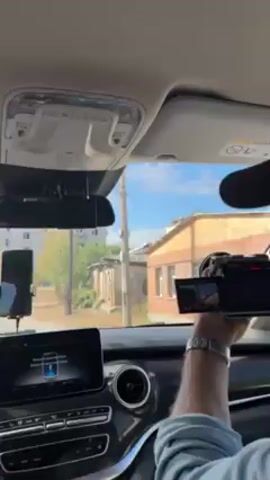 The video shows a person sitting in the passenger seat of a car, facing the camera. They are wearing a black baseball cap, a light blue button-down shirt, and a watch on their left wrist. The person is holding a digital camera with a flip-out screen and appears to be filming out the car window. The camera screen shows the view outside, including buildings and trees. The interior of the car is visible, including the dashboard, steering wheel, and seats. The car is moving, as evidenced by the changing scenery outside the window. The environment outside the car consists of residential buildings and trees, suggesting a suburban or urban area. The sky is blue with some clouds.