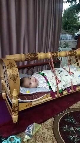A baby is lying in a wooden cradle. The cradle is decorated with intricate carvings and is suspended by ropes. The baby is covered with a blanket adorned with a cartoon pattern. The scene appears to be indoors, with a curtain visible in the background.