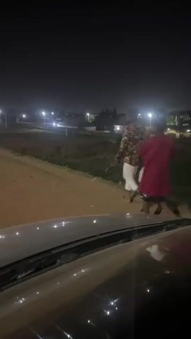 Two individuals walk along a dirt road at night, as seen from the perspective of a vehicle. The first individual is wearing a red top and dark shorts, while the second individual is wearing a patterned shirt and white shorts. They are both walking away from the vehicle, in the same direction. The road is bordered by vegetation. Streetlights illuminate the area. Houses are visible in the background. The hood of the vehicle is visible in the foreground.