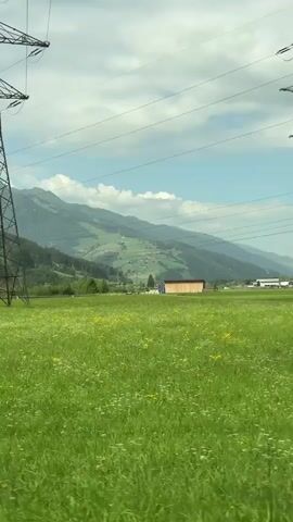 This is a vertical video recording of a passing landscape, likely from a vehicle. The foreground is dominated by a vibrant green field with scattered wildflowers. In the mid-ground, a large transmission tower stands to the left of the frame, with power lines stretching across the sky. Further into the mid-ground, a small, light-colored wooden building is visible. Beyond the field, rolling green hills rise into majestic mountains, some with snow-capped peaks. The sky is a bright blue with scattered white clouds. The camera is moving, as evidenced by the slight blur of the power lines and the changing perspective of the landscape. There are no visible people or animals.