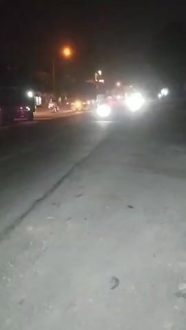 The video shows a blurry, nighttime scene of a road with traffic. Headlights from oncoming vehicles are prominent, creating bright streaks of light. There are streetlights and illuminated signs visible in the background. The road surface appears to be asphalt. A vehicle, possibly a truck, with red taillights is visible on the left side of the frame. The overall impression is a low-light, potentially urban or suburban environment with active traffic.