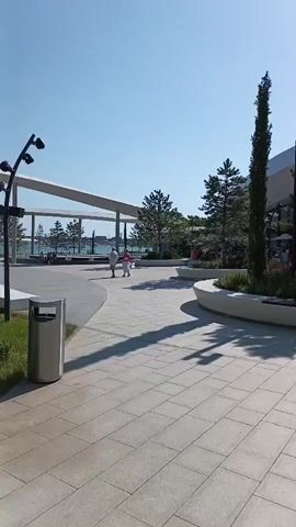 This media file captures an outdoor scene on a sunny day in Neberdzhaevskaya, Russia. The setting appears to be a modern public space, possibly a park or a plaza, characterized by contemporary architecture and landscaping.

In the foreground, a paved walkway with a light-colored, tiled surface stretches across the frame, casting long shadows from trees and structures. To the left, a modern trash receptacle stands near the edge of the walkway.

The midground features several large, curved, white planter beds filled with greenery, including trees and shrubs. Two people, one in a white top and the other in a pink top, are walking in the distance. Behind them, a tall, slender tree stands out, and beyond that, a building with a distinctive, undulating white roof is visible. The building's design is modern and geometric, with large glass windows suggesting a commercial or public facility.

Further into the scene, more trees are scattered throughout the area. The sky is a clear, bright blue, indicating good weather and likely mid-morning or afternoon. Sunlight glints off the building and illuminates the scene.

In the background, the landscape subtly slopes upwards, covered in greenery. Another person is visible walking in the distance near the building. The overall impression is one of a well-maintained, contemporary public area designed for recreation and enjoyment under pleasant weather conditions. There are no discernible emotions conveyed, and no visible text is present in the frame.