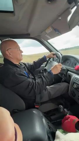 An elderly man with glasses and a bald head is driving a vehicle. He is wearing a black jacket and dark pants. The car's interior is visible, including the dashboard, steering wheel, gear shift, and radio. Outside the car, a rural landscape with fields and trees can be seen. The sky is overcast. There is a red hat and other items on the passenger seat.