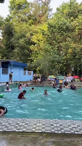 A medium shot captures a bustling swimming pool on a bright day, surrounded by lush green trees. The pool, filled with clear blue water, is alive with activity as numerous children and a few adults splash and play. Several children are seen wearing colorful inflatable rings or tubes, either floating in the water or holding onto them. In the foreground, a young boy is visible, partially submerged in the water, holding a black inflatable tube. To the left of the pool, a bright blue building with white trim is visible, with a person standing near its entrance. Along the edge of the pool, more people are seated, some under colorful umbrellas or canopies, suggesting a recreational area. The overall atmosphere appears lively and joyful, with families and friends enjoying a day at the pool amidst a natural, tree-filled environment. The camera pans slightly to the right, showing more of the pool and the spectators. A woman walks by the edge of the pool, carrying a bag over her shoulder. The lighting suggests it is daytime, possibly late afternoon given the warm tones.