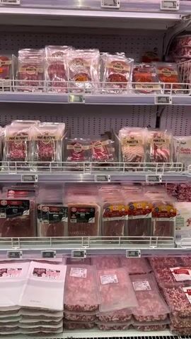 The video pans across shelves of packaged meats in a supermarket. The shelves are well-stocked with various types of cured meats, including ham, bacon, and what appears to be salami or other sausages. The products are predominantly in clear plastic packaging, allowing the contents to be easily visible. Many of the packages have labels with text, though the specific language is not discernible from the video. The lighting is typical of a grocery store, with bright overhead lights illuminating the products. The camera movement is smooth, suggesting a deliberate recording of the display. There are no people visible in the frame. The overall impression is of a well-organized and abundant meat section in a retail setting.