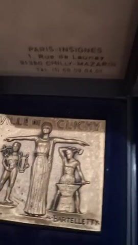 The video shows a close-up of a square, metallic plaque with relief sculptures. The plaque is inscribed with the words "VILLE DE CLICHY" at the top and "BARTELLETTEY" at the bottom. The central figure is a standing woman with her arms outstretched. To her left, a male figure holds a laurel wreath and a lyre. To her right, another male figure is seated at an anvil, holding a hammer, suggesting a blacksmith or industrial theme. The plaque is presented within a dark blue velvet-lined box. The lid of the box, visible at the top of the frame, contains text indicating the manufacturer and address: "PARIS-INSIGNES", "1, Rue de Launey", "91380 CHILLY-MAZARIN", and a phone number. The camera slightly pans and tilts, showing the detail of the relief and the context of the object within its display case.