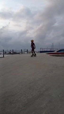 A child wearing a yellow helmet and red shorts and top is skateboarding on a concrete surface at a skatepark. The child rides towards a short metal rail, attempts to ollie onto it, but falls off of the skateboard. Another child walks past in the opposite direction. The skatepark has concrete structures for skateboarding and several palm trees. In the background is a body of water and a cloudy sky.