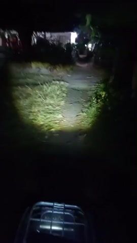 This is a first-person perspective video taken at night from a moving vehicle, likely a bicycle or motorcycle, illuminating a dirt path with a headlight. The path is flanked by vegetation and some buildings are visible in the background, lit by artificial light. The vehicle's dashboard is visible at the bottom of the frame, showing a digital display with the number 15 and the word "GoRider". The video captures the motion of the vehicle as it travels down the path.