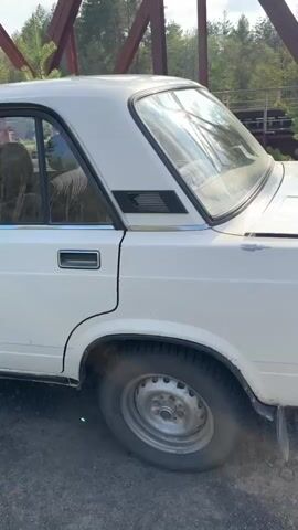 The video pans up from the rear quarter panel of a white 1980s-era Lada 1500 sedan to the top of a brown metal truss bridge. The car is parked on a dirt path next to a forest. The day is sunny with a blue sky and some clouds. A small conifer grows next to the car and a small portion of the bridge’s pedestrian walkway is visible.