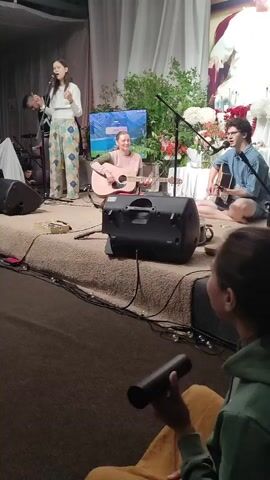 The video shows an indoor live music performance. In the foreground, a woman with dark hair is singing into a microphone while standing. She is wearing a white sweater and patterned pants. To her right, a person with short blonde hair is playing an acoustic guitar and singing. They are wearing a pink and green sweater. Further to the right, a person with dark hair and glasses is also playing an acoustic guitar. They are wearing a blue t-shirt and shorts. Behind the performers, there is a large television screen displaying a nature scene. Plants and flowers are visible, along with stage lighting and audio equipment, including speakers. The stage appears to be carpeted. In the immediate foreground, the back of another person's head is visible, as they hold a black cylindrical object. The overall atmosphere seems to be a casual performance setting.