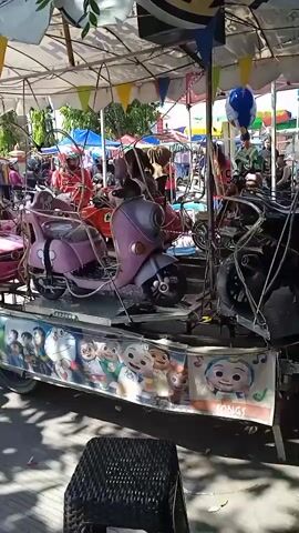 The video shows a children's carousel at an outdoor event, possibly a fair or festival. The carousel is decorated with cartoon characters around its base. It has several ride-on vehicles, including a pink toy car resembling a Mercedes-Benz, a pink scooter, and a green Jeep-style vehicle. There are children riding on the vehicles. The carousel is shaded by a canopy adorned with triangular flags. In the background, people walk around, and there are other tents and stalls, suggesting a bustling atmosphere. A black wicker stool is visible in the foreground. The camera is positioned slightly off-center, offering a view of the carousel as it rotates.