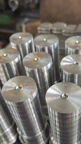 The video displays a close-up, slightly angled shot of multiple cylindrical metal objects with a flared base. These objects are arranged in a clustered formation, with some in the foreground and others receding into the background. Each object features a small, circular indentation at its top surface. The metal appears to be machined, showing faint circular striations. Some of the surfaces exhibit a reddish-brown discoloration, suggesting rust or a similar oxidation. The background is blurred, but it hints at an industrial environment with machinery and other metallic components visible. The lighting is somewhat dim, creating a metallic sheen on the objects. The camera maintains a static position throughout the brief clip.