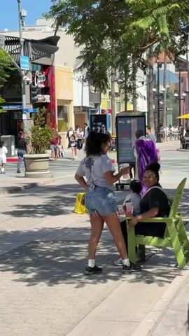 The video shows a sunny outdoor scene with several people. In the foreground, a woman in a grey t-shirt and denim shorts stands holding a drink. To her right, a person in a black t-shirt and a child sit on a green chair. In the middle ground, a man in a plaid shirt and a woman in a green shirt are standing near a bicycle. Further back, two men are seated on colorful chairs under a tree with purple flowers. Across the street, people are walking, and there are buildings and trees. The environment appears to be a pedestrian-friendly street with shops and sidewalks.