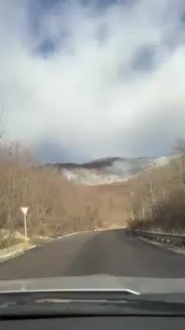 The video depicts a car's point of view driving on an asphalt road heading towards a mountain partially covered in snow. The sky is cloudy with patches of blue. The road is bordered by trees without leaves and a chain-link fence on the right side. There are utility poles and wires running alongside the road. A yield sign is visible at the beginning of the video on the left.