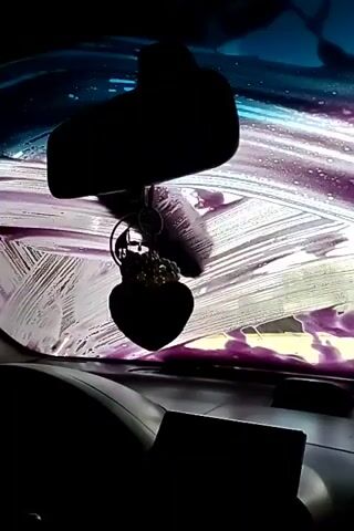 A car is being washed. A rear-view mirror hangs from the ceiling. A black heart-shaped car air freshener with a skull charm hangs from the mirror. The windshield is covered in purple soap suds. The dashboard is visible in the foreground, as well as the steering wheel on the left. The scene is dark.