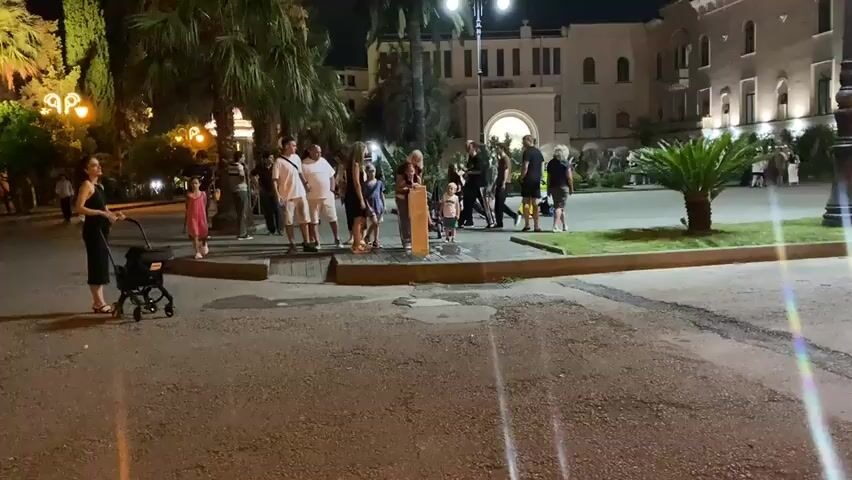 A group of people are gathered around a wooden post in a paved area, possibly a public square or park. Some are standing and observing, while others are interacting with the post. There are children present, some of whom appear to be playing near the post. In the background, there are palm trees and a building with an arched entrance, suggesting an outdoor, possibly tropical or subtropical, location. The lighting indicates it is evening or nighttime. The ground is wet in places, suggesting recent rain or a water feature. People are dressed in casual summer clothing.