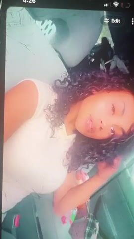 A young woman with curly dark hair and light-colored clothing is in the driver's seat of a car. She is looking at the camera and her lips are parted. The interior of the car is visible, including the dashboard and the side window. Outside the window, trees and a building are visible. The video is shaky and appears to be taken with a phone.