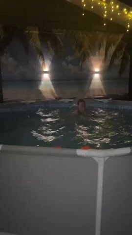 The video shows a girl swimming in a pool outdoors at night. The pool is surrounded by what appears to be a partially enclosed structure, decorated with palm tree images and string lights. Two spotlights illuminate the scene from behind the girl. The girl is wearing a swimsuit and is actively moving around in the water. There is also an orange float in the pool. The overall atmosphere is dark but illuminated by artificial lighting, creating a relaxed and potentially festive mood.