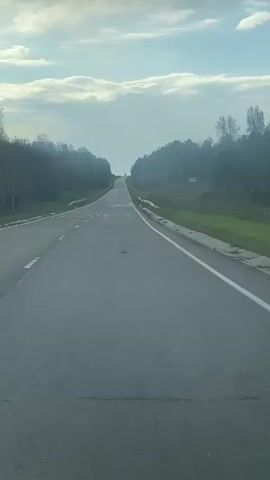 The video shows a perspective from inside a vehicle traveling on a paved road. The road curves gently to the right and extends uphill towards the horizon. Trees line both sides of the road, with a dense forest on the left and sparser trees on the right. The sky is partly cloudy with a bright horizon. The road surface appears to be in fair condition with some visible patches and markings. There are no other vehicles or people visible in the immediate vicinity.