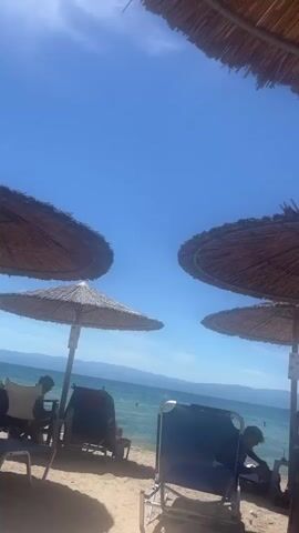 The video shows a beach scene with several straw umbrellas providing shade. There are beach chairs in the foreground. In the background, there is a body of water with distant land visible on the horizon under a clear blue sky. A few people are visible sitting on the chairs or walking on the beach. The camera appears to be moving slightly, possibly handheld.