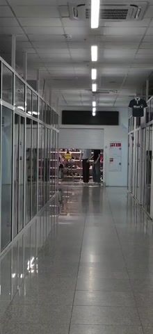 The video shows a long, indoor hallway with shops on either side. The hallway has a polished concrete floor and fluorescent lighting. On the left, glass-fronted shops display merchandise. On the right, a shop has its metal security gate partially rolled up, revealing shelves of shoes and bags. Two mannequins stand in front of this shop, one male and one female, dressed in casual clothing. A dark blue shirt with a logo is displayed on a hook above the shop on the right. The video appears to be shot from a static position at the end of the hallway, looking towards the partially opened shop.