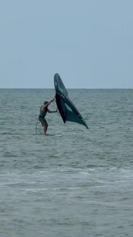 A person is wing foiling on a calm body of water. The person is standing on a white board and holding a large, teal and black wing above their head. The wing is angled to the right and appears to be catching the wind. The water is a muted blue-gray, with small waves on the surface. The sky is overcast and a pale gray. The horizon is visible in the distance, where the water meets the sky. The person is wearing a dark tank top and shorts, and a life vest is visible. The scene is shot from a medium distance, with the person and their equipment as the central focus.