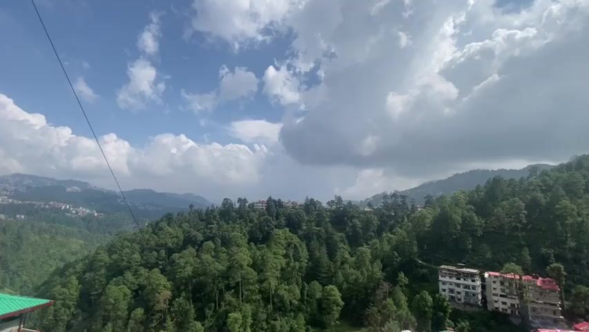 A wide shot captures a dense forest on a mountainside under a partially cloudy sky. In the foreground, a single dark wire stretches across the top left of the frame. The mountains are covered in numerous tall, green pine trees, with a few buildings visible nestled among them on the slopes. On the right side of the frame, a multi-story building with light-colored walls and dark windows is situated on the hillside. The sky above is a mix of blue and white, with large, fluffy clouds. The overall scene depicts a natural landscape with human habitation integrated into the mountainous terrain.