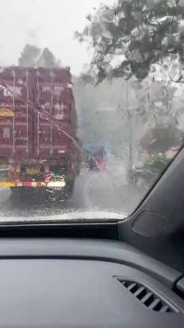 The video is filmed from inside a vehicle looking out through a rain-streaked windshield. The view shows a highway in what appears to be heavy rain, indicated by the water droplets on the windshield and the blurred, wet appearance of the road and surrounding trees. Traffic is visible ahead, including a large red shipping container truck in the left lane and other vehicles further down the road. The interior of the car is partially visible, showing a dashboard and speaker grill. The overall impression is one of a difficult drive in adverse weather conditions.