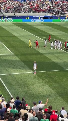 This is an aerial, long shot of a soccer game. The field is green and striped with white lines. A player in a white and blue uniform stands in the foreground, looking toward the goal. In the distance, players in red and white uniforms are lined up near the goal. A goalie in a yellow uniform stands in the goal. Spectators are visible in the stands in the background. The crowd is large and the stadium is open-air. Advertising boards are visible around the stadium.