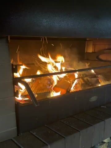 The video shows a close-up view of a fire burning in a fireplace. Logs are stacked and burning with bright orange flames and wisps of smoke. Sparks are seen flying upwards from the fire. The fireplace appears to be made of brick and metal. The lighting is dim, highlighting the flames and the warm glow of the fire. There are no people or other objects visible in the frame. The overall scene conveys a sense of warmth and coziness.