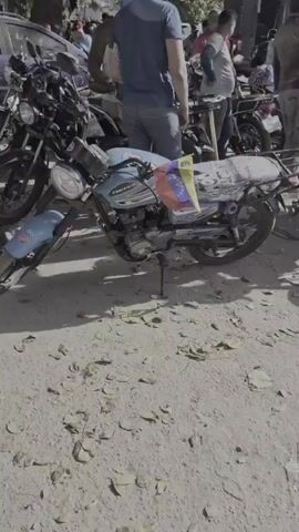The video shows a group of people gathered around motorcycles in an outdoor setting. In the background, a damaged building or pile of rubble is visible, suggesting a recent disaster or construction site. Some individuals are on or near motorcycles, while others are standing and talking. The lighting indicates it is daytime. One motorcycle prominently displays a Venezuelan flag. The overall scene appears to be chaotic or in the aftermath of an event, with people seeming to be navigating the environment, possibly for transportation or to assess the situation.