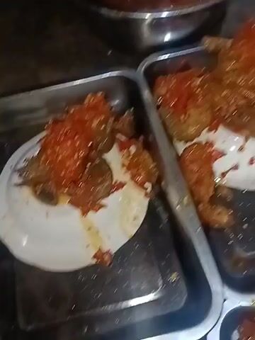A person is pouring a red sauce from a large metal bowl into two rectangular metal trays. The trays contain food items, possibly chicken or meat, which are covered in the red sauce. The sauce appears to be chunky, with visible pieces of chili or other vegetables. The environment looks like a kitchen or food preparation area, with various objects and surfaces visible in the background, including a stovetop and cleaning supplies. The lighting is dim, and the video quality is low, making it difficult to discern finer details.