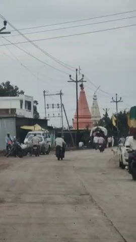 The video depicts a street scene in what appears to be a rural or semi-urban area. In the background, two temple spires are visible, one predominantly orange-pink and the other white. Power lines crisscross the sky, supported by utility poles. On the left side of the street, a white building with a corrugated metal roof extension is present. Several motorcycles and a white van are parked near this building. A group of people are gathered, some standing and others on motorcycles or a bicycle. In the foreground, the street is unpaved and dusty. Several individuals are riding motorcycles down the street, and one person is riding a bicycle. Two dogs are also visible on the right side of the street. The overall atmosphere suggests a typical day with people moving about their daily activities, possibly a market or gathering near the temples. The sky is overcast, indicating a cloudy day.