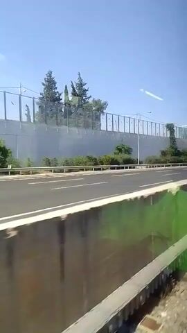This video shows a view from a moving vehicle, likely a train, looking out at a highway. The highway has multiple lanes and is separated by white lines. A concrete barrier with a fence on top runs along the side of the highway, presumably to dampen noise. Trees are visible behind the barrier. The camera angle suggests the viewer is traveling at a considerable speed. The time stamp indicates the video is very short, lasting only 5 seconds.