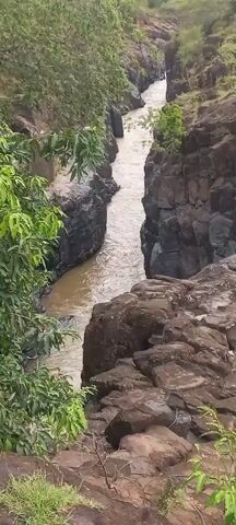 A river flows through a narrow canyon. The water is murky brown. The canyon walls are made of dark rock with some sparse vegetation. The camera is positioned on top of a rock formation, looking down into the canyon.
